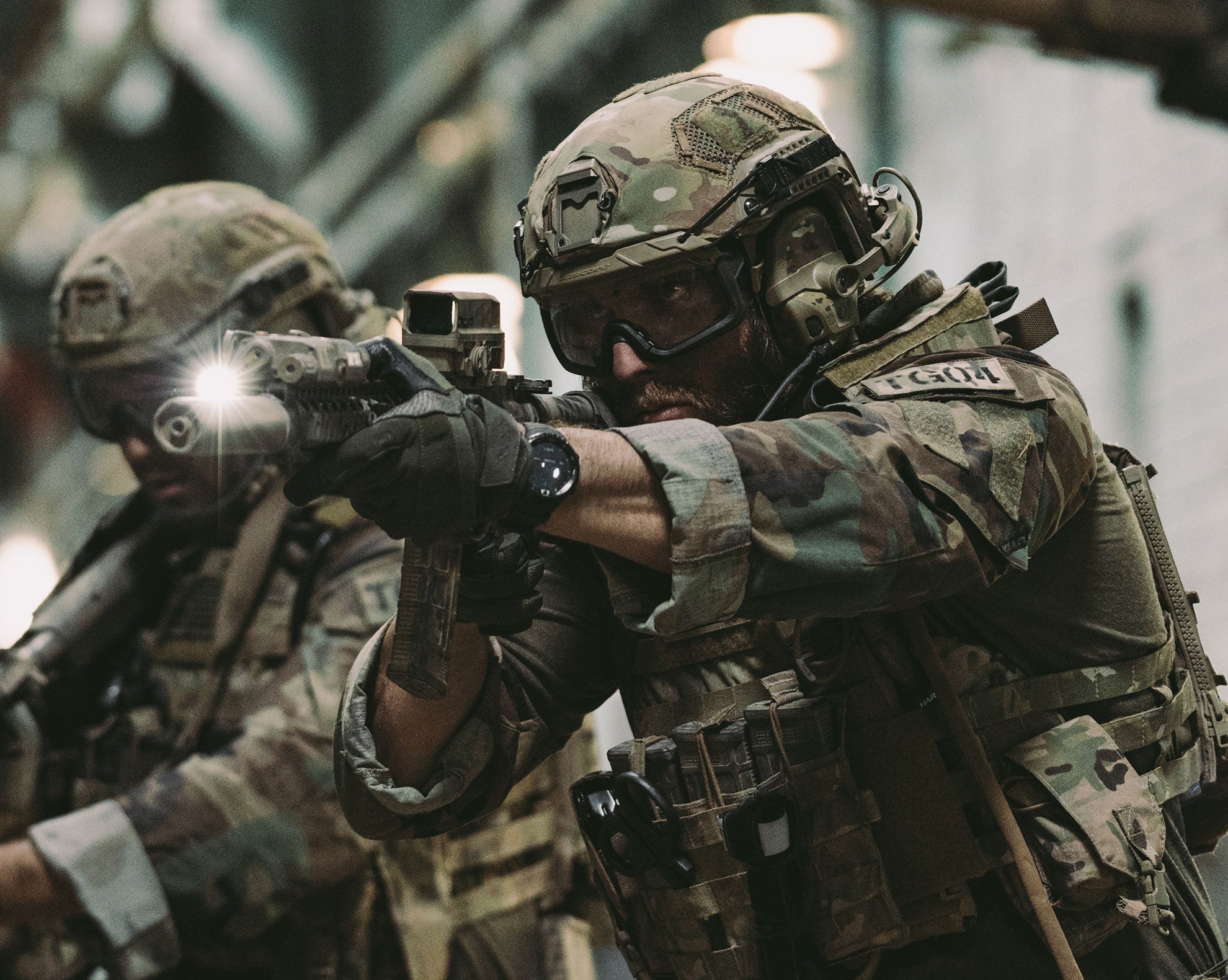 Two soldiers in camouflage gear with one aiming a rifle.